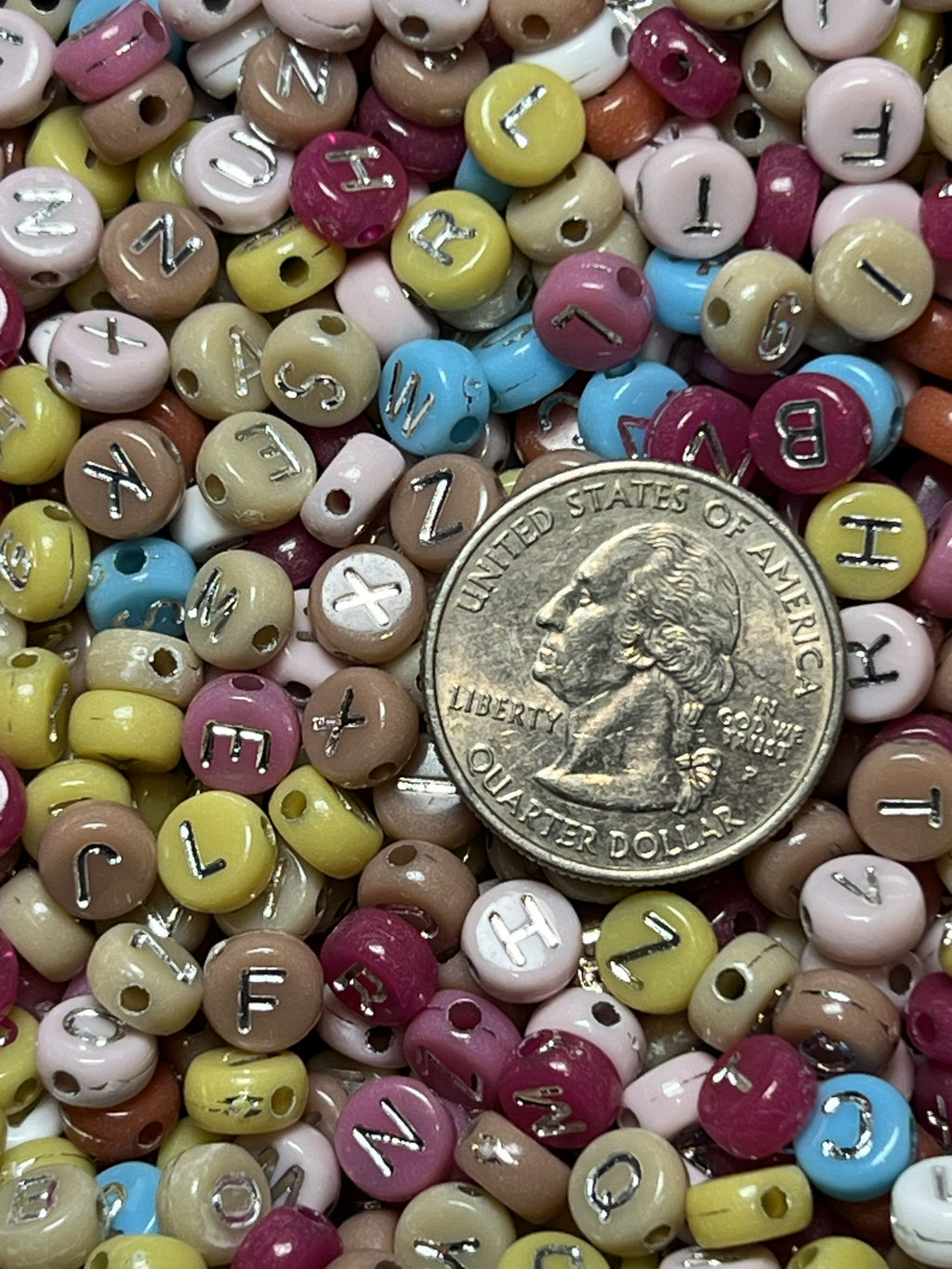 A pile of colorful acrylic beads with silver letters, shown next to a US quarter for scale.