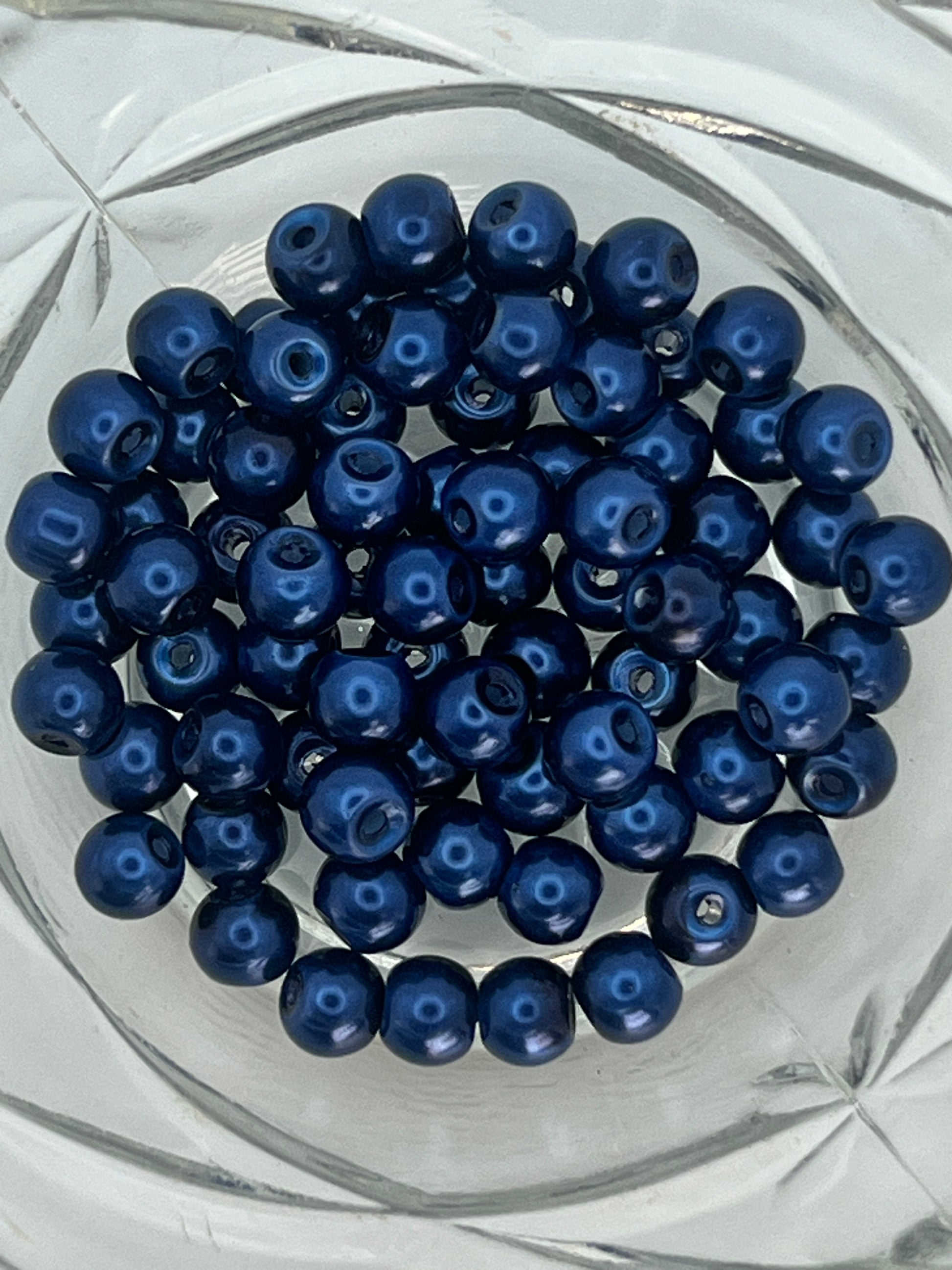 A collection of 6mm steel blue glass pearl beads in a clear glass bowl.