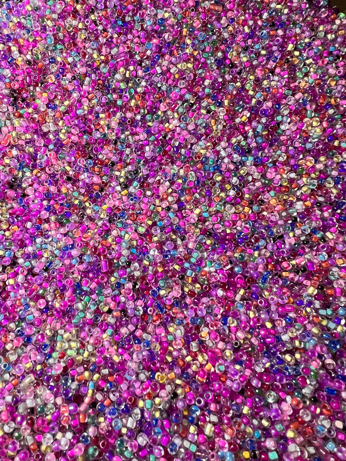 A dense pile of mixed 8/0 glass seed beads in vibrant shades of pink, purple, and blue.